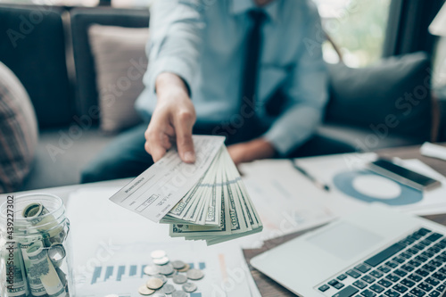 A Businessperson's hand giving cheque and dollar bill to customer and coin, laptop and graph chart on the desk at office. Payment by check, paycheck, payroll concept.