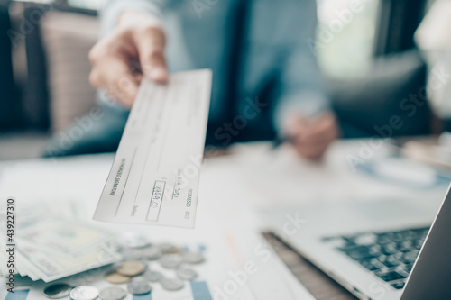 A Businessperson's hand giving cheque to customer and dollar bill, coin, laptop and graph chart on the desk at office. Payment by check, paycheck, payroll concept.