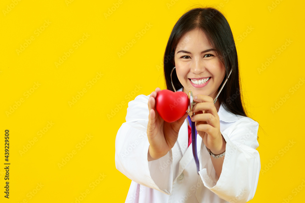 Asian cheerful female doctor wearing white gown uniform, using stethoscope to check up at mini heart with isolated yellow background and copy space. Medical treatment, Disease and Healthy concept. Asian cheerful female doctor wearing white gown uniform, using stethoscope to check up at mini heart with isolated yellow background and copy space. Medical treatment, Disease and Healthy concept.
