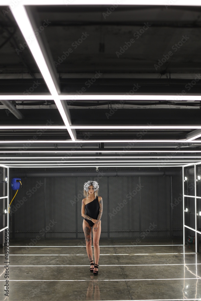 A girl in a black bodysuit with bare legs and special foil on her head stands indoors where there are many artificial fixtures. Fashion model. High quality photo
