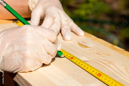 Close-up. Female hands in rubber light gloves hold a green pencil and a yellow tape measure on a wooden board for measuring dimensions. 