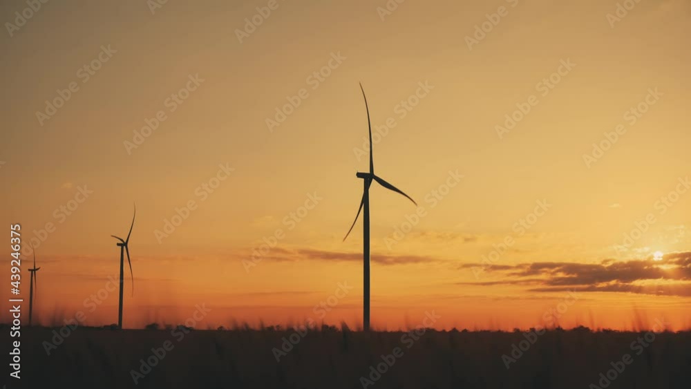 Silhouettes of wind turbines with rotating propellers producing electricity in field against orange sky at summer sunset Silhouettes of wind turbines with rotating propellers producing electricity in field against orange sky at summer sunset