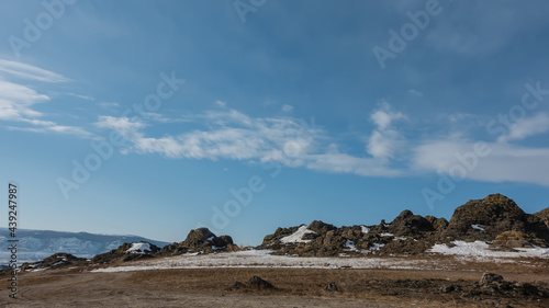 Winter landscape. There is snow and dry grass in the valley. Against the background of the blue sky, there is a mountain range devoid of vegetation. Picturesque clouds. Siberia