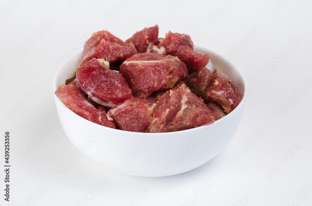 Ceramic bowl with raw marinated meat on a white background. Pieces of pork with salt, pepper and granulated garlic.