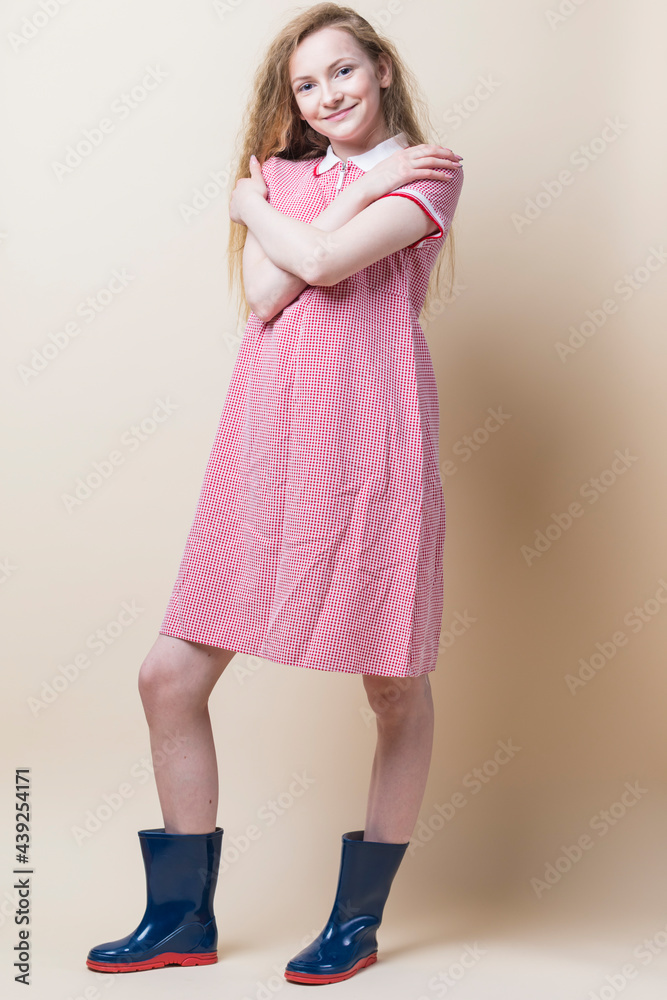 Full Length Portrait of Smiling Caucasian Teenager Girl In Red Checked Dress and Wellington Boots Posing With Crossed Hands Over Beige.
