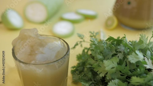 A slice of juicy lemon falling inside a glass of refreshing bottle gourd squash - Giya juice. Closeup shot of a Lauki cut into half  green Dhania  and nutritious drink against a yellow background photo
