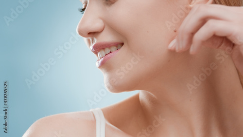 Young slim good-looking European woman with long golden hair in white bikini with her head turned smiles wide for the camera touching her jawline against blue ripple background | Skincare concept