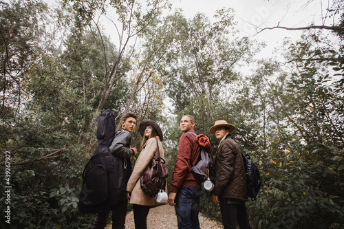 Four young people in warm clothes with guitar and backpacks standing in grove with green trees and looking back