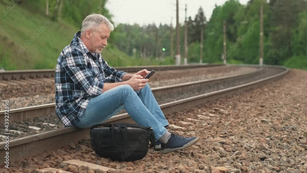 An elderly male tourist is sitting on the railway tracks, looking through information on the phone and waiting for the train. A middle-aged tourist waits for a train on the railway on a summer day. An elderly male tourist is sitting on the railway tracks, looking through information on the phone and waiting for the train. A middle-aged tourist waits for a train on the railway on a summer day.
