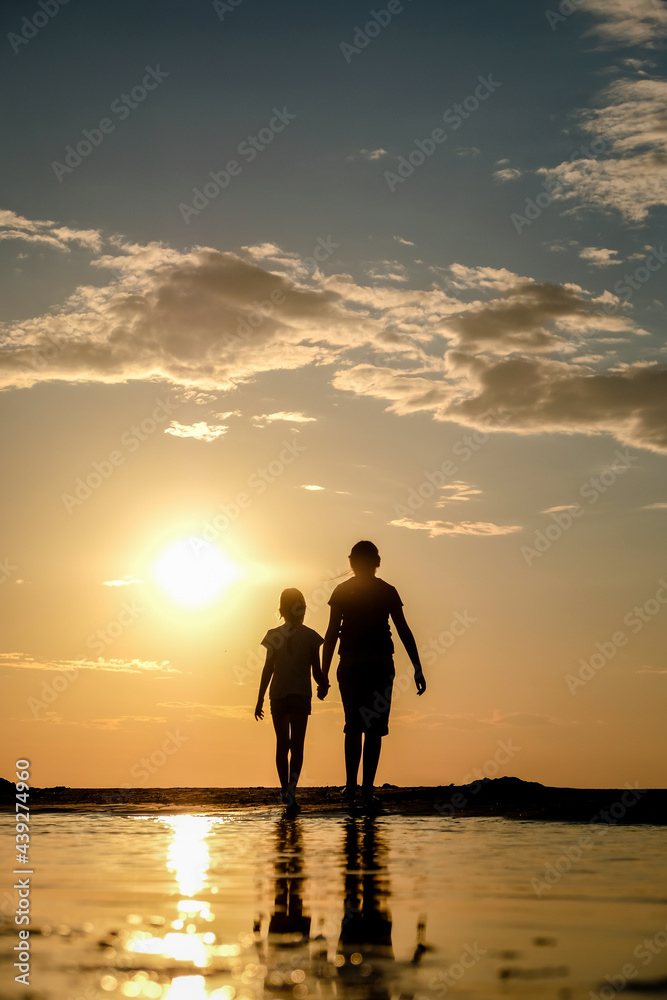 Silhouette of two sisters holding hands who standing on the beach at the sunset. Vertical photo