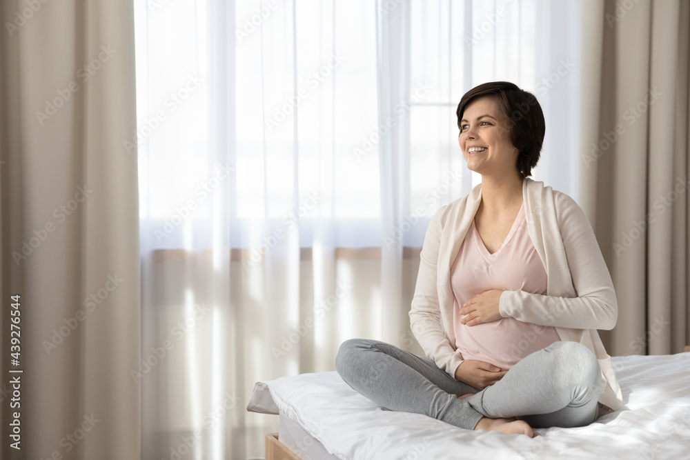 Smiling dreamy pregnant woman touching belly, sitting on cozy bed at home, happy young future mom expecting first baby, relaxing in bedroom, enjoying healthy pregnancy, motherhood concept