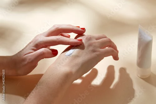 Obraz beauty product, cosmetics and people concept - female hands with tube of cream applying moisturizer to skin on beige background