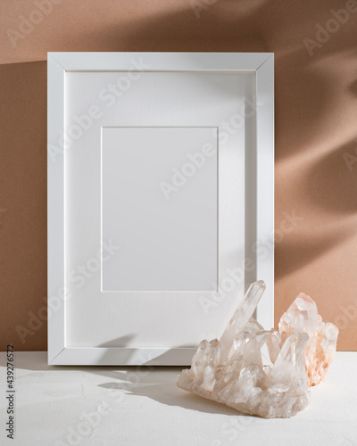 Empty white frame with mat, natural crystal, illuminated by sunlight. Poster mock up with a place for your design in a nude interior.