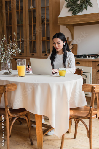 serious young asian woman in earphones sitting on table  using laptop and writing with pen in notebook in kitchen