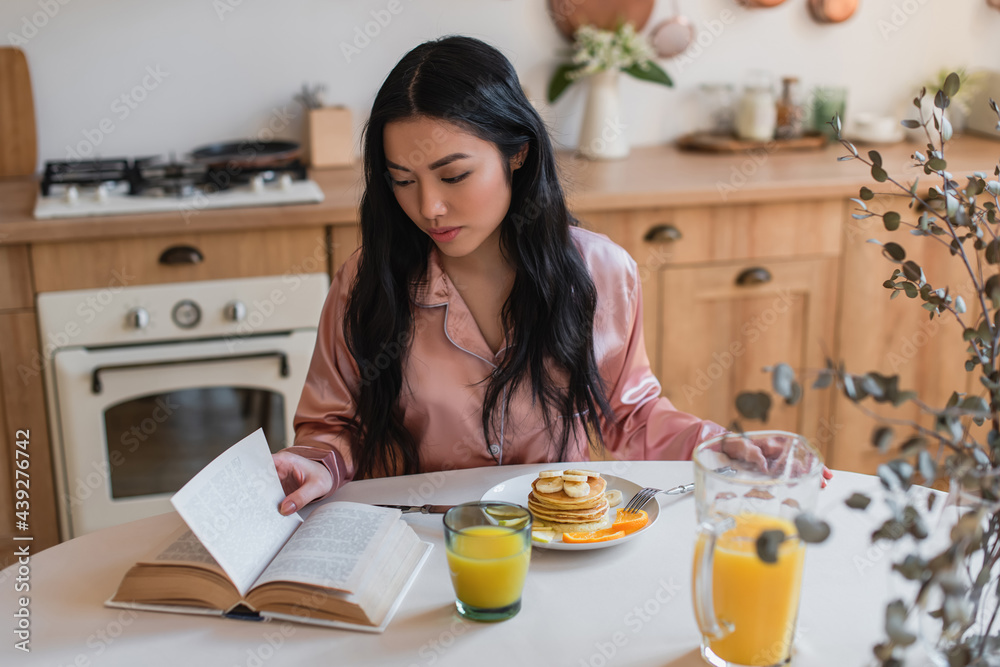 young asian girl in silk pajamas reading book while having delicious breakfast in kitchen young asian girl in silk pajamas reading book while having delicious breakfast in kitchen