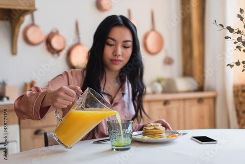 young asian woman in silk pajamas pouring orange juice into glass near pancakes and cellphone on table in kitchen