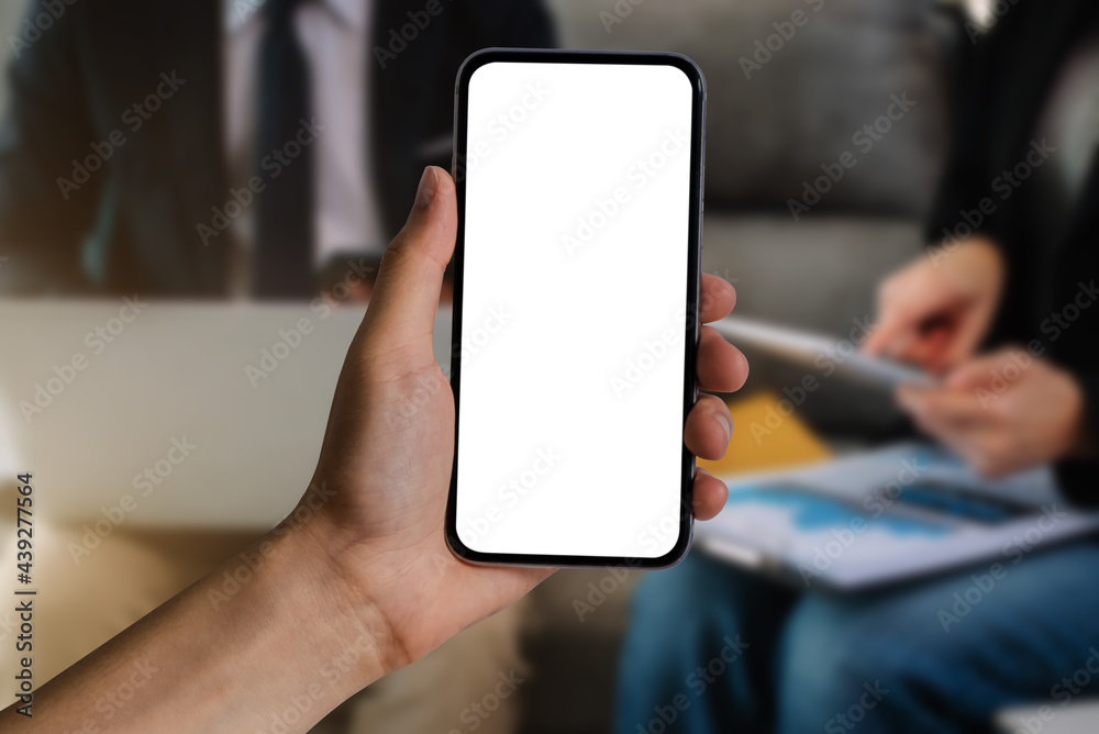 Hand holding white mobile phone with blank white screen in office.