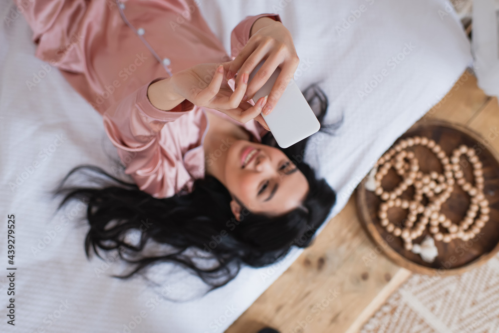 top view of smiling young asian woman in silk pajamas lying on bed and taking selfie with cellphone in bedroom
