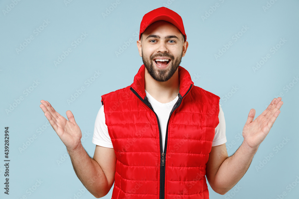 Professional delivery guy employee man in red cap white T-shirt vest uniform workwear work as dealer courier spreading hands isolated on pastel blue color background studio portrait. Service concept. Professional delivery guy employee man in red cap white T-shirt vest uniform workwear work as dealer courier spreading hands isolated on pastel blue color background studio portrait. Service concept.
