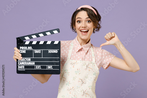 Young housewife housekeeper chef cook baker woman wear pink apron hold classic black film making clapperboard poitn thumb finger on herself isolated on pastel violet background Cooking food concept. photo