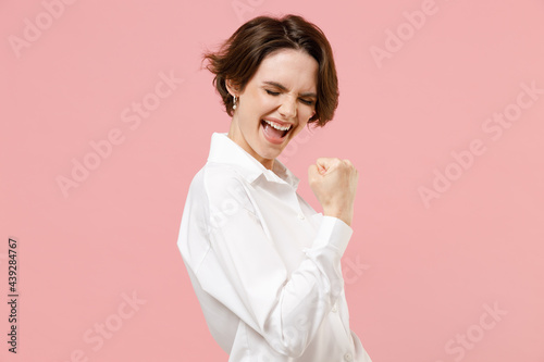 Young fun overjoyed successful employee business woman corporate lawyer in classic formal white shirt work in office do winner gesture clench fist say yes isolated on pastel pink background studio. photo