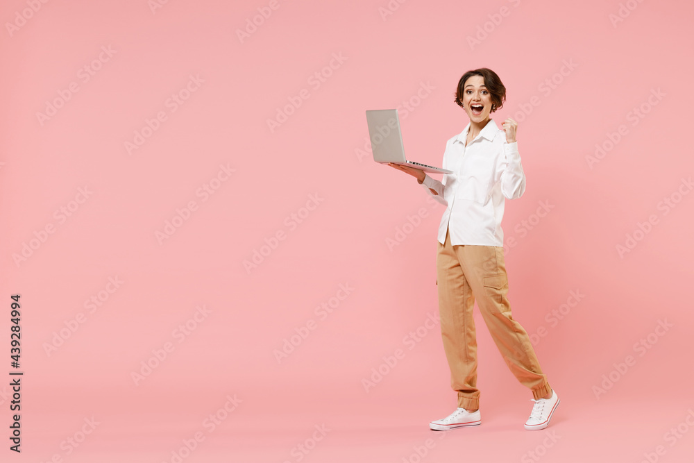 Full length young employee business woman corporate lawyer in classic white shirt work in office hold laptop pc computer chat online do winner gesture clench fist isolated on pastel pink background Full length young employee business woman corporate lawyer in classic white shirt work in office hold laptop pc computer chat online do winner gesture clench fist isolated on pastel pink background