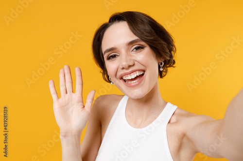 Close up young smiling happy fun friendly woman 20s with bob haircut wearing white tank top shirt doing selfie shot on mobile phone waving hand isolated on yellow color background studio portrait