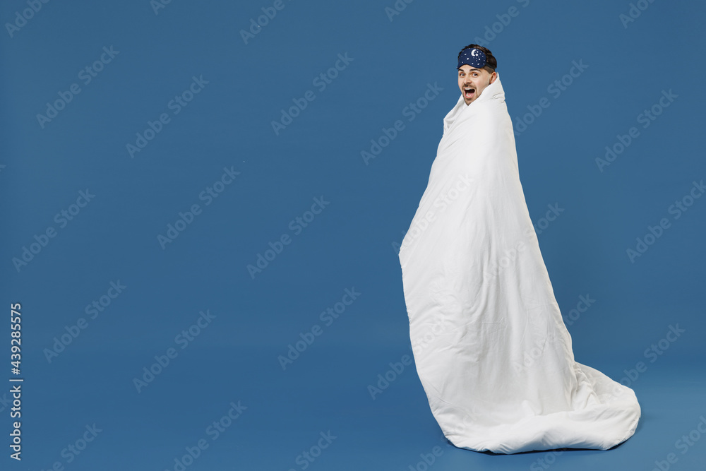 Full length side view young surprise man 20s in pajamas jam sleep mask resting relax at home wrap cover with blanket duvet look camera isolated on dark blue background Good mood night bedtime concept