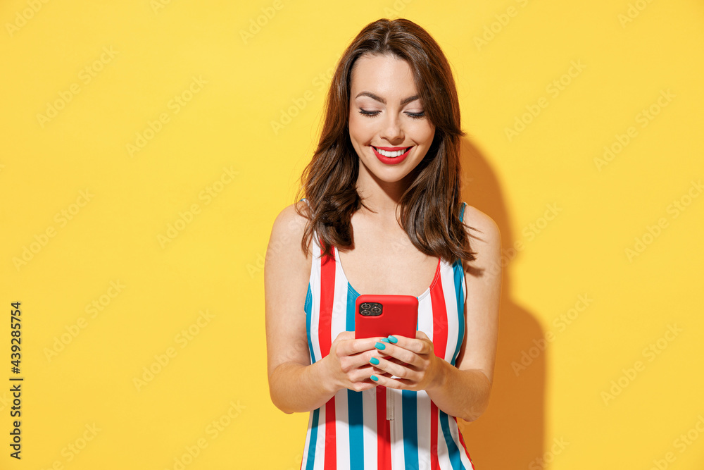 Happy young sexy woman slim body wear striped red blue swimsuit hold using mobile cell phone send sms isolated on vivid yellow color wall background studio. Summer hotel pool sea rest sun tan concept. Happy young sexy woman slim body wear striped red blue swimsuit hold using mobile cell phone send sms isolated on vivid yellow color wall background studio. Summer hotel pool sea rest sun tan concept.