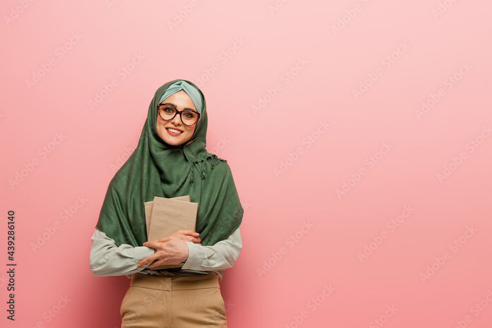 Fototapeta premium pretty muslim teacher with textbooks smiling at camera on pink background