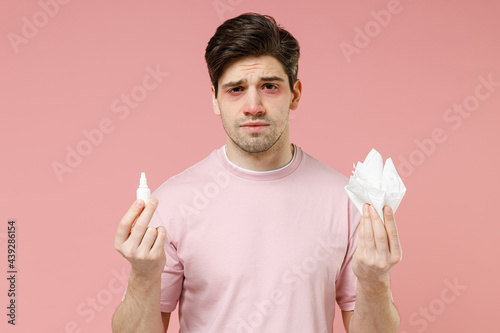 Frustrated sick unhealthy ill allergic man has red eyes runny stuffy sore nose suffer from allergy symptoms hay fever hold paper napkin use nasal drops isolated on pastel pink color background studio
