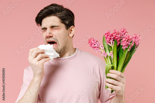 Fototapeta Sick allergic man has red eyes runny stuffy sore nose suffer from pollen allergy symptoms hay fever hold bloom flower plant napkin reaction on trigger isolated on pastel pink color background studio