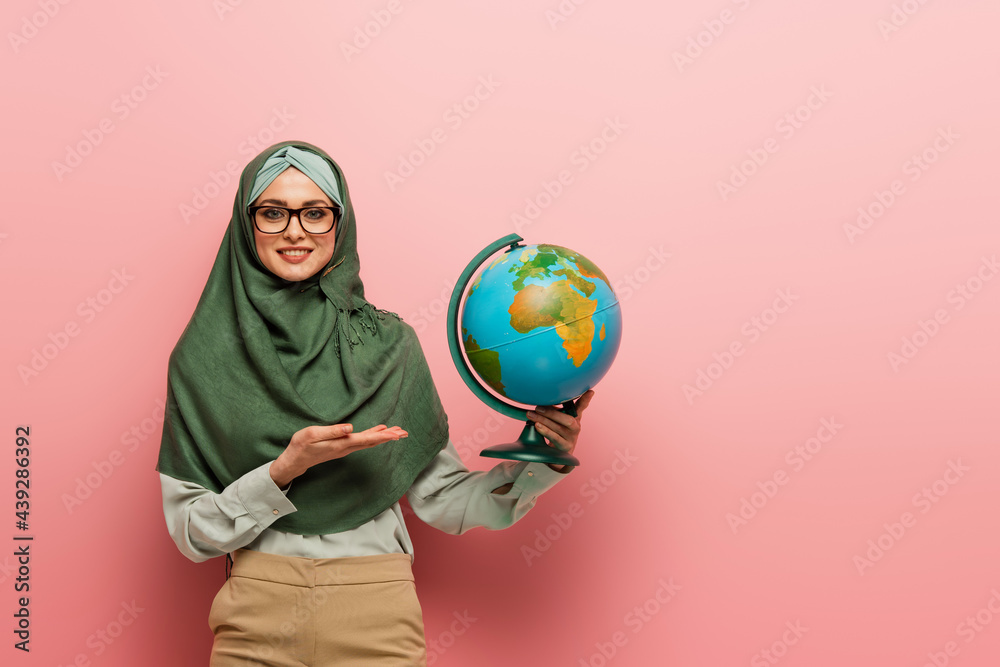 smiling muslim teacher in green hijab pointing at globe on pink background smiling muslim teacher in green hijab pointing at globe on pink background