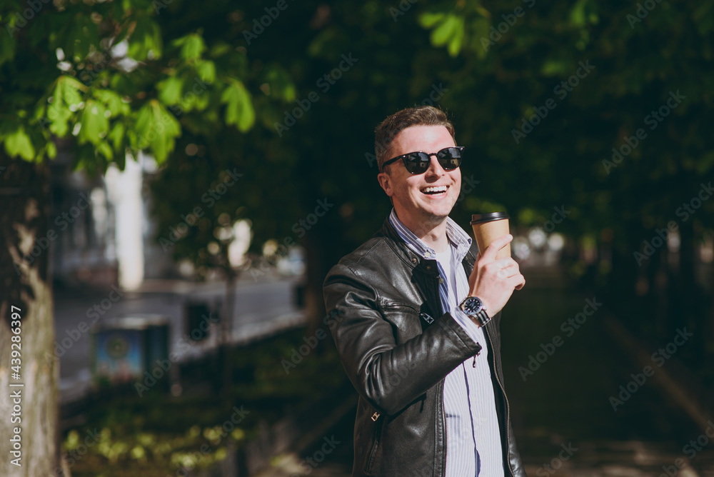 Young european happy smiling man in black leather jacket eyeglasses strolling in green park hold takeaway delivery craft paper cup drink coffee to go. Concept of people urban lifestyle spring weather. Young european happy smiling man in black leather jacket eyeglasses strolling in green park hold takeaway delivery craft paper cup drink coffee to go. Concept of people urban lifestyle spring weather.