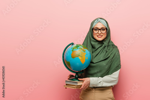 smiling muslim teacher with textbooks and globe looking at camera on pink background photo