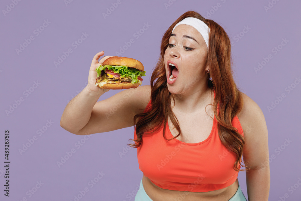 Young happy fun chubby overweight plus size big fat fit woman wear red top warm up training hold fast food eating bite burger isolated on purple background home gym Workout sport motivation concept Young happy fun chubby overweight plus size big fat fit woman wear red top warm up training hold fast food eating bite burger isolated on purple background home gym Workout sport motivation concept