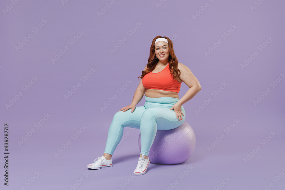 Full length side view young chubby overweight plus size big fat fit woman wear red top warm up training sit akimbo on fit ball rest isolated on purple background gym. Workout sport motivation concept. Full length side view young chubby overweight plus size big fat fit woman wear red top warm up training sit akimbo on fit ball rest isolated on purple background gym. Workout sport motivation concept.