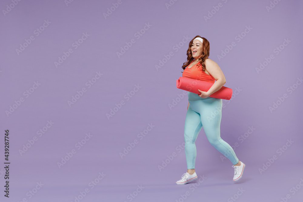 Full length side view happy young chubby overweight plus size big fat woman 20s wear red top warm up train hold yoga mat walk isolated on purple background gym home. Workout motivation sport concept Full length side view happy young chubby overweight plus size big fat woman 20s wear red top warm up train hold yoga mat walk isolated on purple background gym home. Workout motivation sport concept
