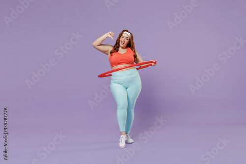 Fototapeta Naklejka Na Ścianę i Meble -  Full length young chubby overweight plus size big fat woman wear red top warm up train twisting hula hup point thumb finger on herself isolated on purple background gym home. Workout sport concept