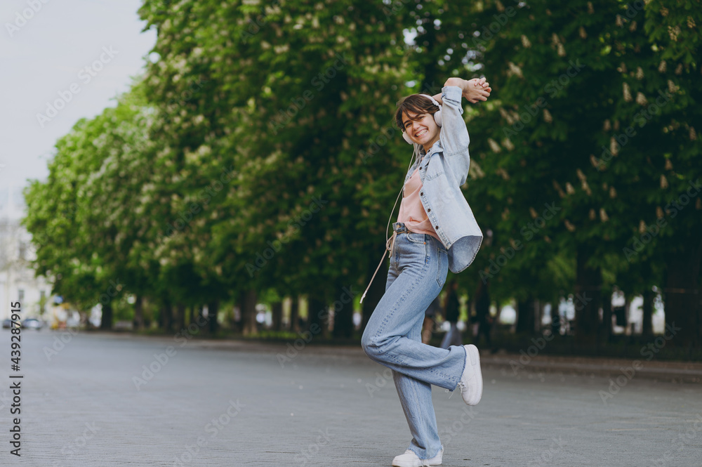 Full length side view young woman in jeans clothes headphones listen to music walking strolling down green park alley outdoors dancing hold raised up hands above head People urban lifestyle concept