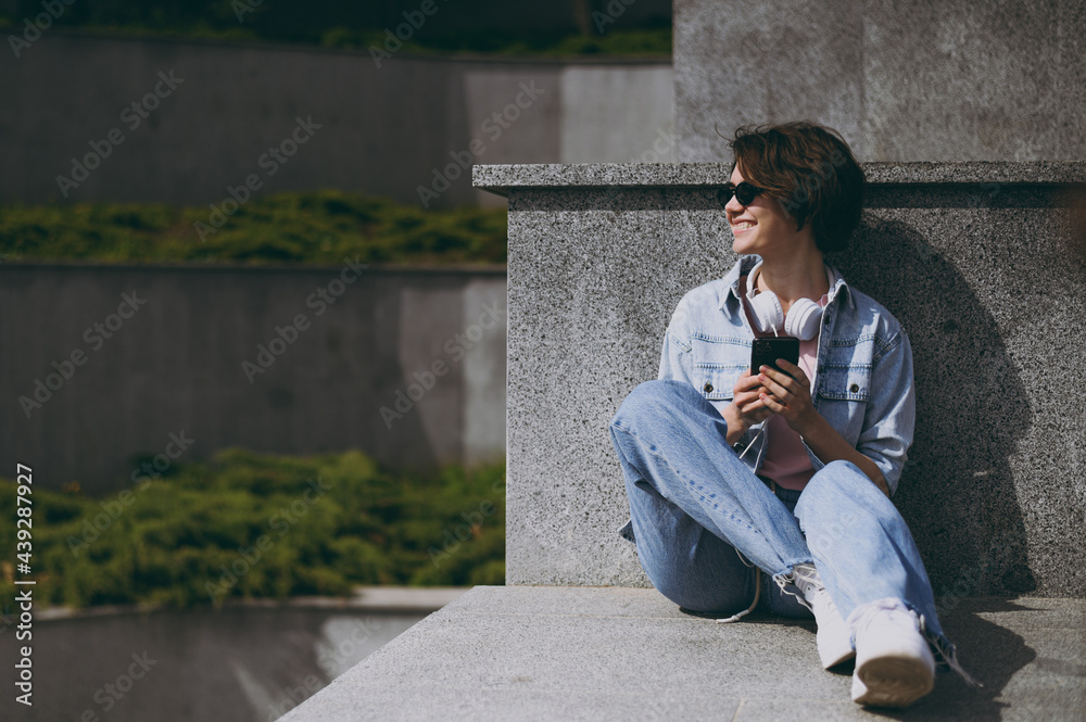 Full length young student woman in jeans clothes headphones glasses listen to music leaning on building wall sit on concrete steps outdoors use mobile cell phone look aside People lifestyle concept.