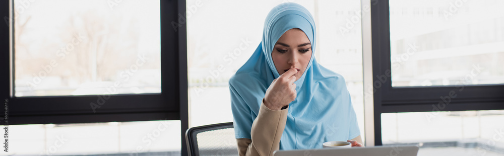 Fototapeta premium arabian businesswoman in hijab holding coffee cup while thinking against window, banner