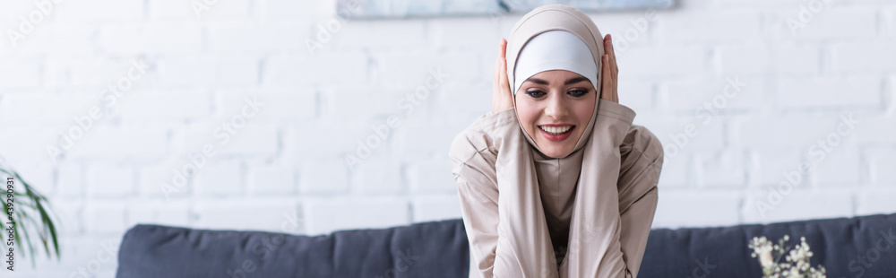 cheerful muslim woman touching head while smiling at home, banner cheerful muslim woman touching head while smiling at home, banner