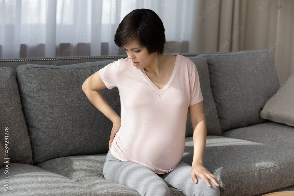 Tired pregnant woman touching back, massaging tensed muscles, sitting on couch at home, exhausted young future mom feeling pain from baby bump or backache, health problem and pregnancy concept