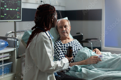 Senior man breathing with help from oxygen tube laying in hospital bed, listening african american doctor explaying diagnosis treatmant holding pills bottle for recovery. photo