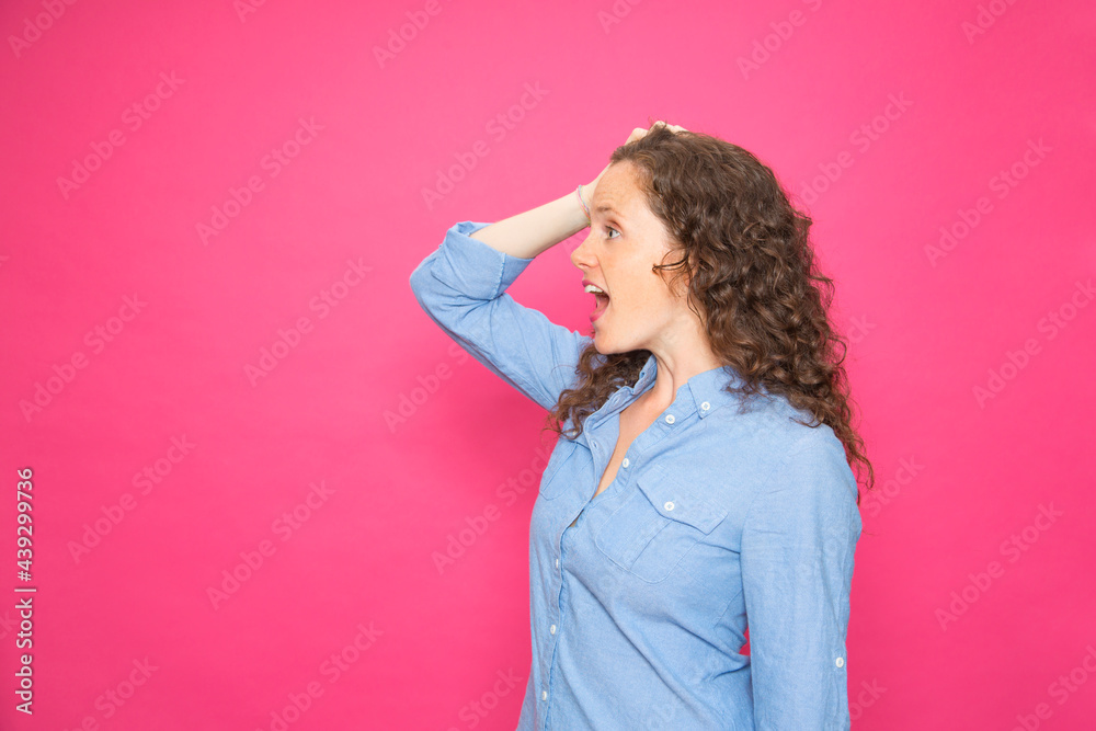 Portrait happy young pretty girl sideways with hand on head screaming surprised in blue shirt on pink background Portrait happy young pretty girl sideways with hand on head screaming surprised in blue shirt on pink background