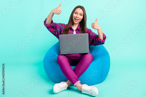 Portrait of trendy beautiful cheerful girl sitting in chair using laptop showing thumbup isolated over bright teal color background © deagreez