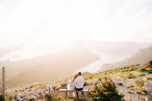 The bride and groom are tenderly embracing and sitting on a wooden bench in the mountains overlooking the Bay of Kotor 