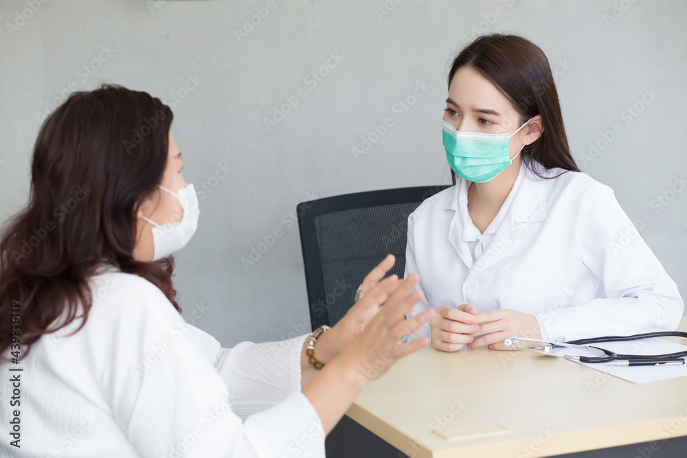 An Asian female patient is attentively explaining her condition to a professional female doctor in the clinic, both wearing masks to prevent infection at the hospital. An Asian female patient is attentively explaining her condition to a professional female doctor in the clinic, both wearing masks to prevent infection at the hospital.