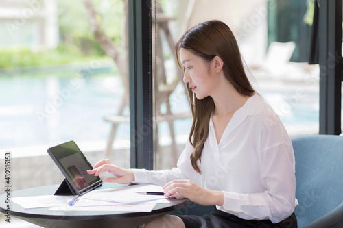 Asian confident woman wear a white shirt is using a tablet to work and there are many documents on the table. her face with smiling in a working room at home.